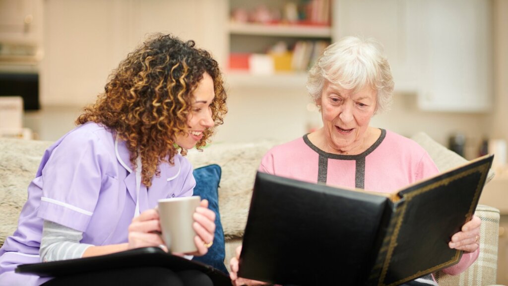 An elderly woman and caregiver sitting on a couch together looking at a photo book and a caregiver holding a coffee mug