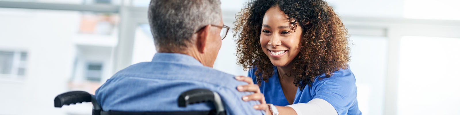 A senior man sitting in a wheelchair as his nursing assistant looks at him with her hand on his shoulder