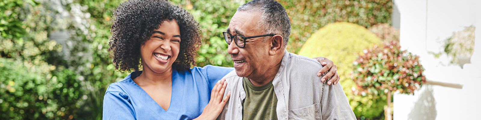 A senior man walking outdoors as his nursing assistant puts her arms around his shoulders to help steady him